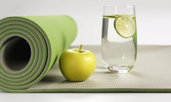 Yoga fitness, health life style. Yoga rolled green mat , green apple, glass of water with lemon on white background. Place for text. Healthy lifestyle, fitness, sport concept