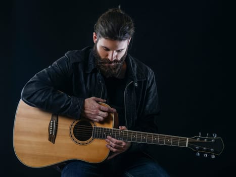 Young bearded man sitting and playing an acoustic guitar over black background.