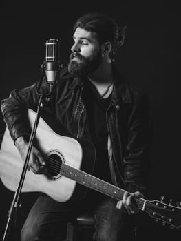 Young bearded man sitting, playing an acoustic guitar and singing over black background.
