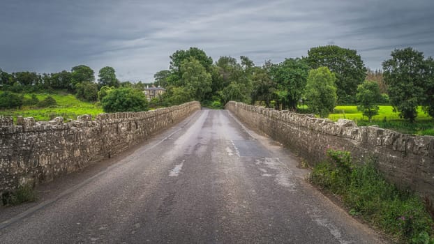 Road leading over old, 12th century stone Bective Bridge over Boyne River with stone wall on sides, Count Meath, Ireland