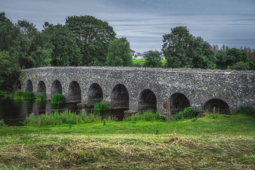 Closeup on old, 12th century stone arch Bective Bridge over Boyne River surrounded by green fields and forest, Count Meath, Ireland