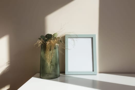 Glass vase with herbarium flowers, eucalyptus branches, empty white photo frames on the white table in the interior with beige walls near window.