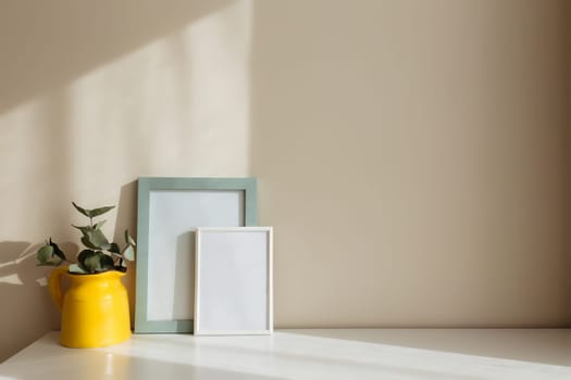 A yellow ceramic jug or vase with eucalyptus branches, empty white photo frames on the white table in the interior with beige walls near window.