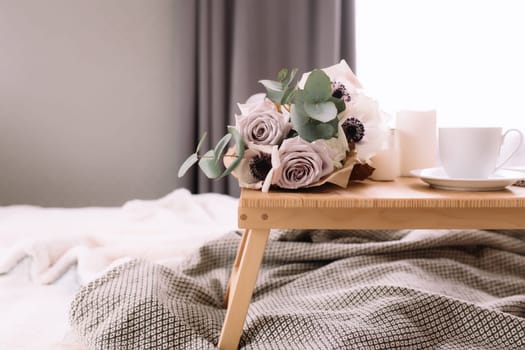 Romantic morning. Wooden coffee table with flowers on bed with plaid, coffee cup, flowers and candles. Lilac roses with eucalyptus and anemones. Interior gray tones. High quality photo