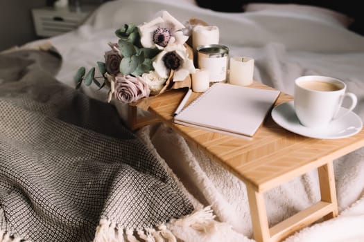 Romantic morning. Wooden coffee table with flowers on bed with plaid, coffee cup, flowers and candles, note pad, pen. Lilac roses with eucalyptus and anemones. Interior gray tones. High quality photo