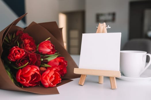 A bouquet of red tulips on the table and a white cup of coffee, empty white frame. Concept for a greeting card. High quality photo