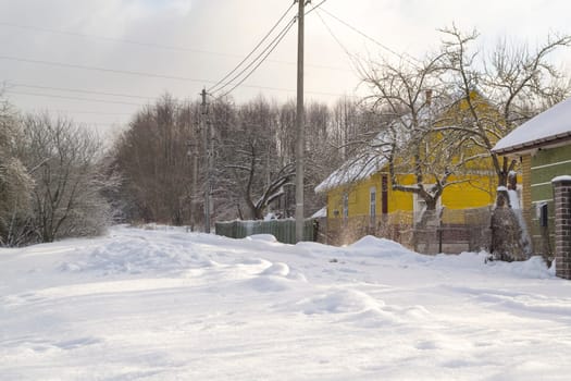 A cloudy winter day with snowfall. A street in a dacha village. Snow covered rural road and snowdrifts along the road near the fences in Belarus