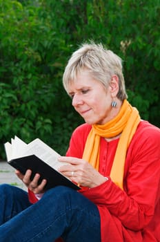 Side view of a mature woman sitting at the edge of a park and looking into an open book that is being held in front of her.