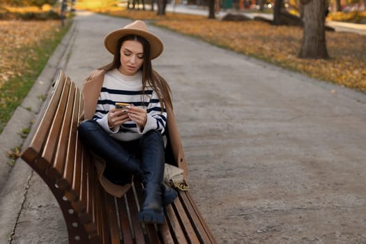 Attractive cheerful brunette girl in trench coat with coffee to go happily using cellphone outdoor.