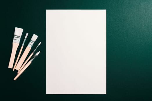 A clean white sheet and paint brushes on a dark green background with space to copy. Mock-up, mockup, layout.