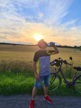 Handsome young professional cyclist dressed in cycling clothing and protective helmet feeling free and happy, drinking water from the bottle, enjoying bicycle in warm spring. Recreation of sportsman.