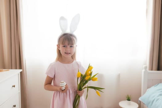 Easter concept. Small smiling girl in white fluffy bunny ears and pink dress holding yellow tulip flowers and colorful egg. Room interior. High quality photo