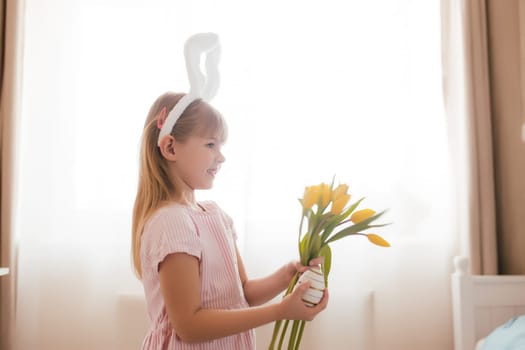Easter concept. Small smiling girl in white fluffy bunny ears and pink dress holding yellow tulip flowers and colorful egg. Room interior. High quality photo