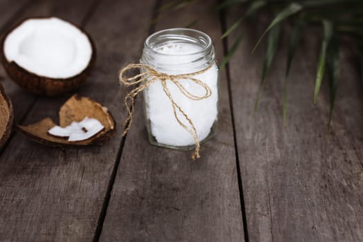 Broken coconuts on gray wooden background with jar of raw organic extra virgin coconut oil and palm leaf. White coconut pulp. High quality photo