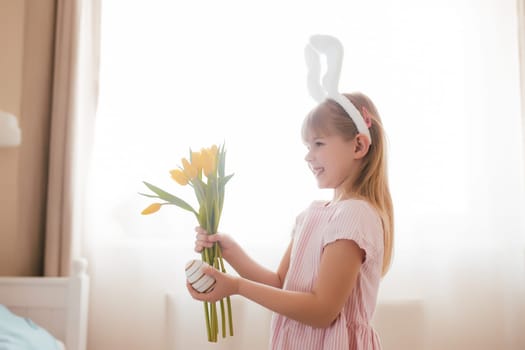 Easter concept. Small smiling girl in white fluffy bunny ears and pink dress holding yellow tulip flowers and colorful egg. Room interior. High quality photo