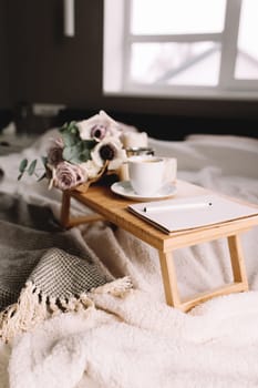 Romantic morning. Wooden coffee table with flowers on bed with plaid, coffee cup, flowers and candles, note pad, pen. Lilac roses with eucalyptus and anemones. Interior gray tones. High quality photo
