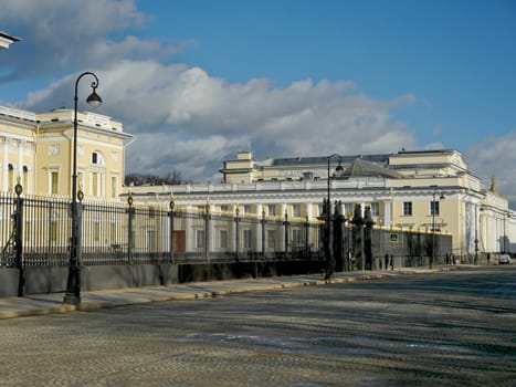 Ancient palace behind a high cast-iron fence, lonely passers-by walk along the cobbled pavement, the panorama of the old city, a sunny spring day, cumulus clouds are flying across the sky