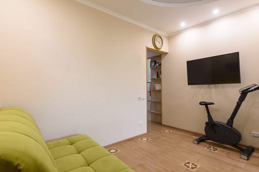 Fragment of the living room interior, in the foreground there is a sofa, on the back wall there is a TV and an exercise bike