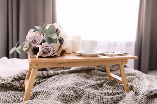 Romantic morning. Wooden coffee table with flowers on bed with plaid, coffee cup, flowers and candles, note pad, pen. Lilac roses with eucalyptus and anemones. Interior gray tones. High quality photo