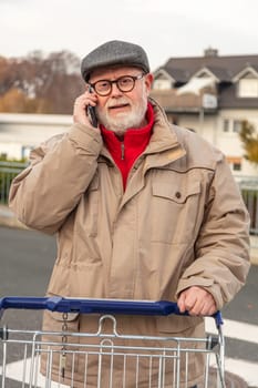Senior with winter cloth using cell phone while he is pushing is shopping cart on parking area