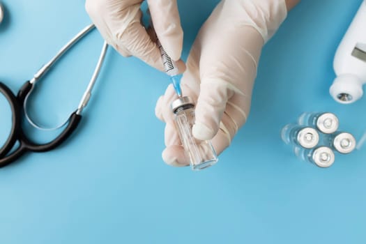 Medicine treatment, vaccination concept. Doctor hands holding bottle with vaccine and injection syringe, electronical thermometer, stethoscope and vaccine vial on blue background. High quality photo