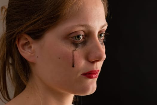 Young woman crying in depression from profile on black background. Blonde female with blurry maskara in pain. Unhappy girl with blank look in darkess.