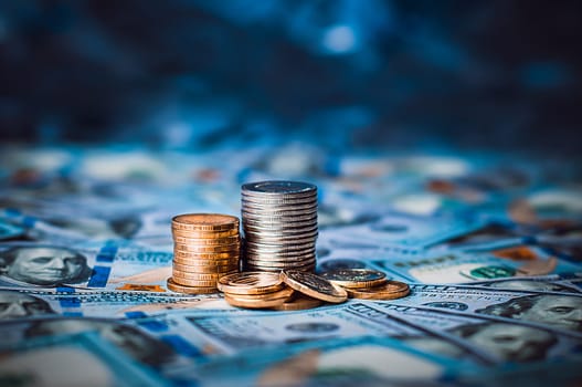 Stacks of coins against the background of hundred-dollar bills are scattered throughout the space. The coins are collected in two columns, they are shiny, golden in color.