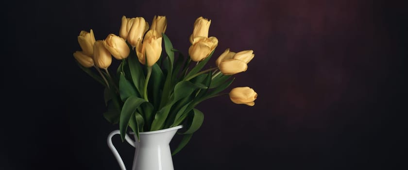 Tulips in a jug. Classic still life with a bouquet of yellow tulip flowers in a vintage white jug on a dark background and an old wooden table. Art photography.
