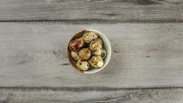 Table top view - small ceramic bowl full of spotted quail eggs on gray wood desk.
