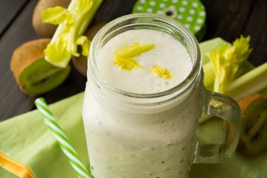 green smoothie in a glass jar with lid and a straw, celery and kiwi over dark wooden table
