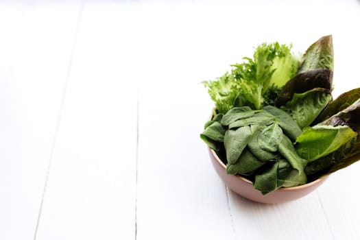 Flat lay with bowl of fresh green salad leaves of spinach and lettuce, romaine and basil on white wooden background. Healthy vegetarian eating concept. High quality photo