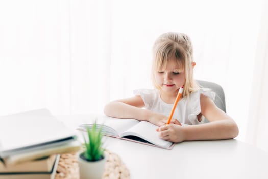 A little girl sits at the table and writes in a white notebook. Education concept. Home schooling. Homework. High quality photo