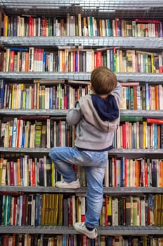 Badajoz, Spain - October 18th, 2020: Child boy climbing bookshelves at second hand book bookshop. Encourage good reading habits in kids