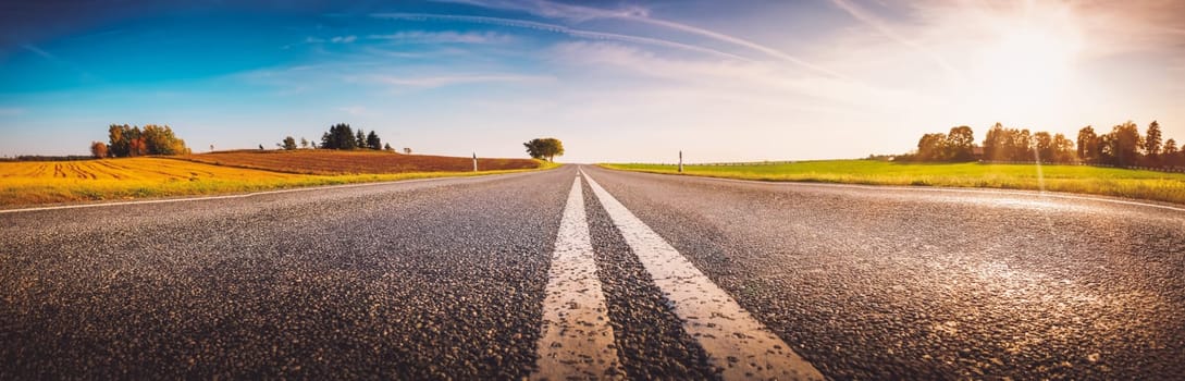 Wide asphalt road panorama in countryside in sunset on summer. Route with white dividing lines in beautiful nature landscape.
