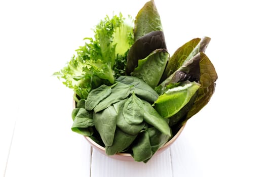 Flat lay with bowl of fresh green salad leaves of spinach and lettuce, romaine and basil on white wooden background. Healthy vegetarian eating concept. High quality photo