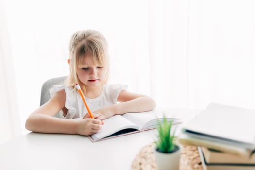 A little girl sits at the table and writes in a white notebook. Education concept. Home schooling. Homework. High quality photo