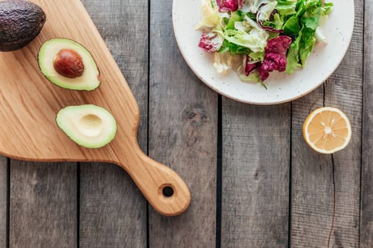 Healthy eating concept flat lay. Mediterranean diet, plate with fresh green lettuce salad leaves, purple radicchio leaves, cutting board with avocado halves, kernel, lemon on wooden background