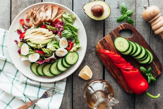 Healthy eating concept flat lay. Mediterranean diet, plate with lettuce salad leaves, quail egg, avocado, tomato, cutting board with cucumber, pepper, basil, olive oil, lemon on wooden background