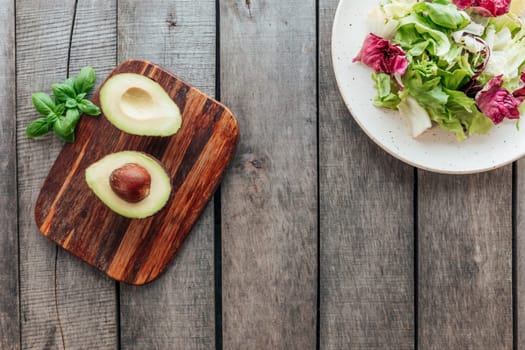Healthy eating concept flat lay. Mediterranean diet, plate with fresh green lettuce salad leaves, purple radicchio leaves, cutting board with avocado halves, kernel, basil bunch on wooden background