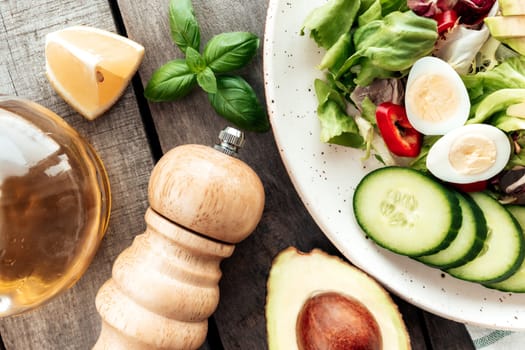 Healthy eating concept flat lay. Mediterranean diet, plate with lettuce salad leaves, quail eggs, avocado half with kernel, cucumber slices, pepper, basil, olive oil, lemon, salt on wooden background