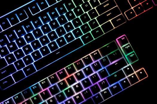 top view detail closeup of two illuminated mechanical gaming RGB keyboard on black desk next to each other with keycaps lighting up in different colors in the dark