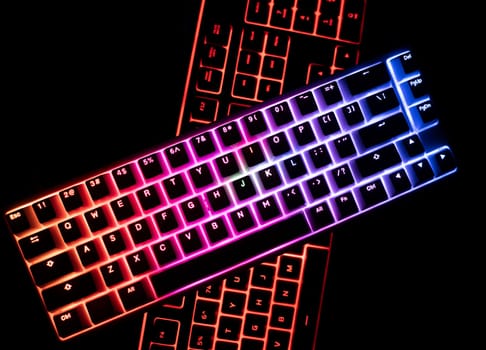 top view detail closeup of two illuminated mechanical gaming RGB keyboard on black desk on top of each other with keycaps lighting up in different colors in the dark