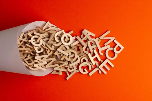 top view closeup of small wooden alphabet letters getting spilled from a white paper cone on orange background