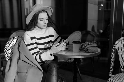 beautiful smiling woman in stylish outfit sitting at the table in a hat and sweater, romantic happy mood, waiting for a boyfriend on a date in a cafe, spring-summer fashion trend, drinking coffee.
