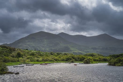Panoramic view on mountains of Killarney National Park with wooden boats on Lough Leane and dark dramatic clouds in background, Ring of Kerry, Ireland
