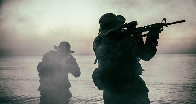 Commando soldiers walking in water, army special operations forces fighters sneaking in darkness, aiming assault rifles and observing shore during amphibious operation on coast at night or dawn