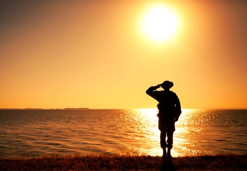 Silhouette of saluting commando soldier, army infantryman standing on shore during sunset or sunrise. Military solemn ceremony, respectable salute for honoring fallen heroes and comrades veterans