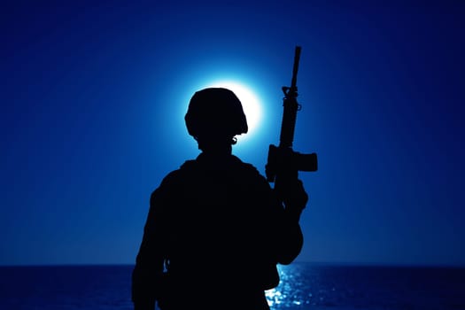 Silhouette of army soldier standing with service rifle in hand on background of night sky with moon. Armed Marines infantry in battle helmet and uniform ready for action, patrolling area in darkness
