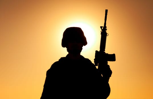 Silhouette of army soldier, special operations forces infantry rifleman, Marines fighter in combat helmet and ammunition standing with assault rifle in hand on background of sunset or dawn sky