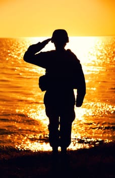 Silhouette of army soldier, Marines fighter in helmet and ammunition saluting while standing on seashore at sunset time. Coast guard fighter, special operations forces soldier honoring fallen comrades